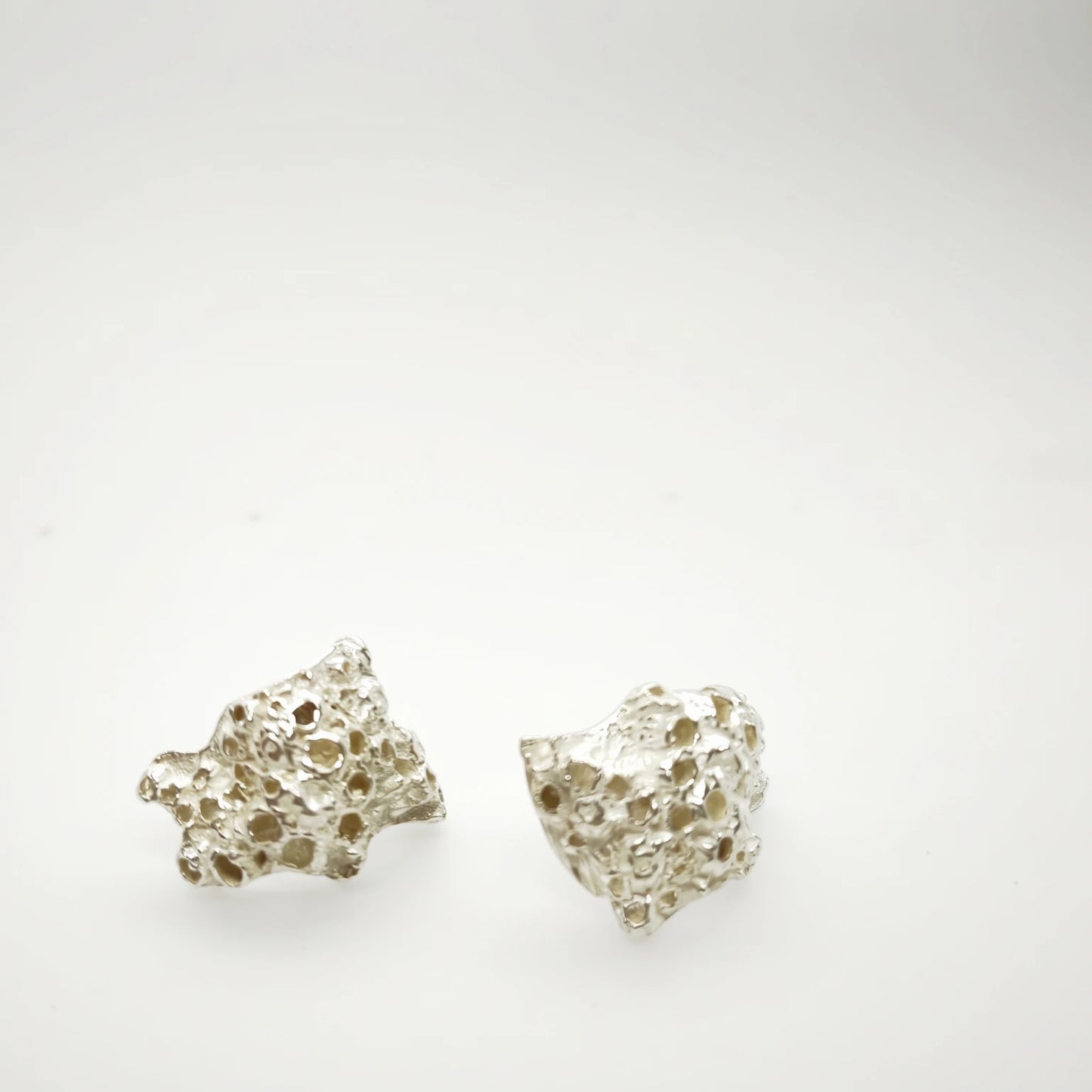 A pair of textured, organic-shaped silver earrings with a porous, porous-like surface, displayed on a clean white background.