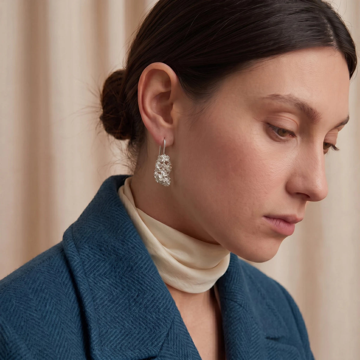 A close-up of a woman wearing a textured silver earring with clear stones, paired with a cream turtleneck and a blue herringbone blazer, set against a soft beige curtain background.