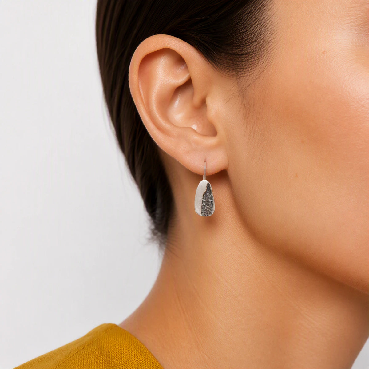 Sterling silver drop earring featuring a black bottle motif worn on the ear, shown in a close-up contemporary jewellery portrait against a soft white background, by Inês Almeida.
