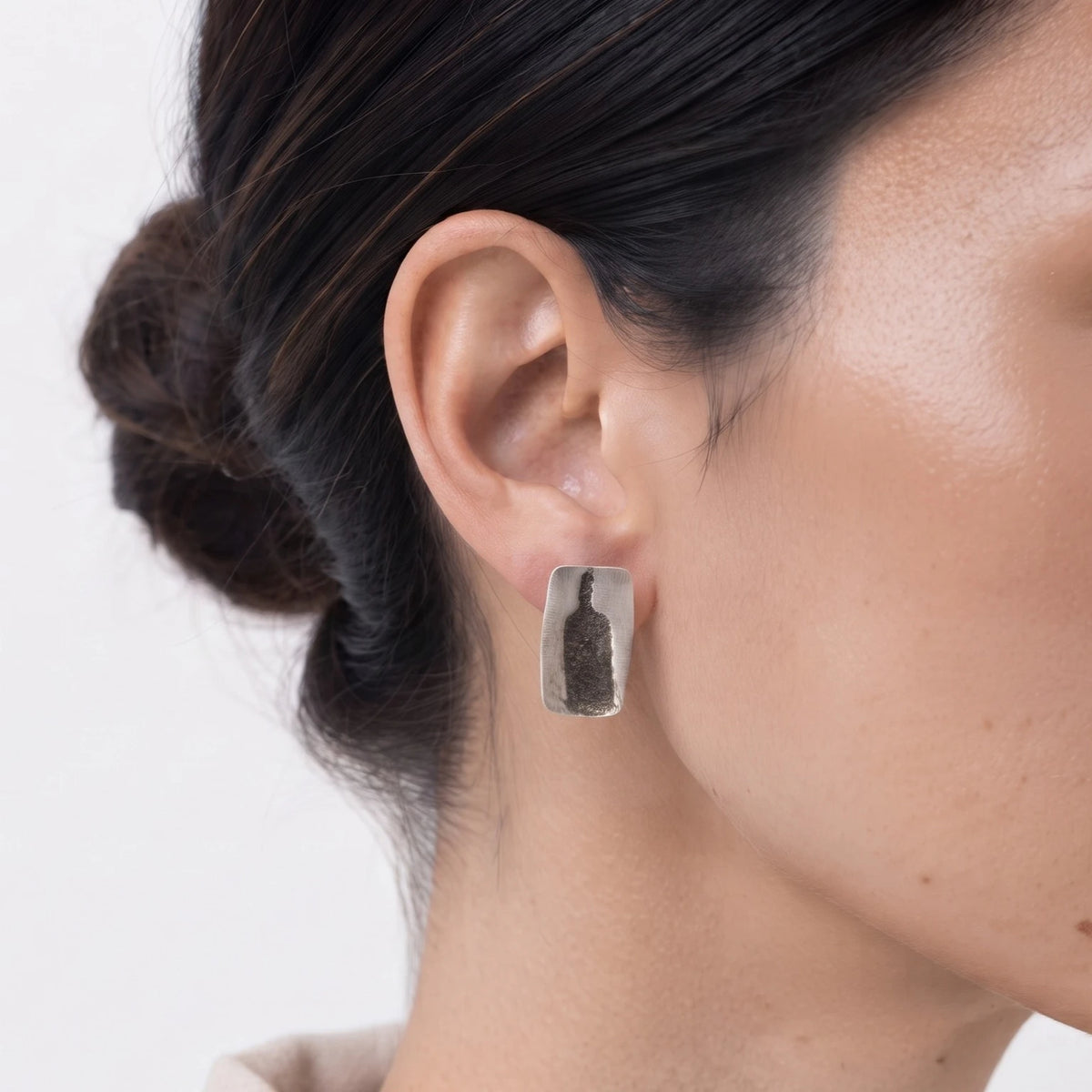 Sterling silver earrings featuring a bottle motif worn on the ear, shown in a close-up contemporary jewellery portrait against a soft white background, by Inês Almeida.