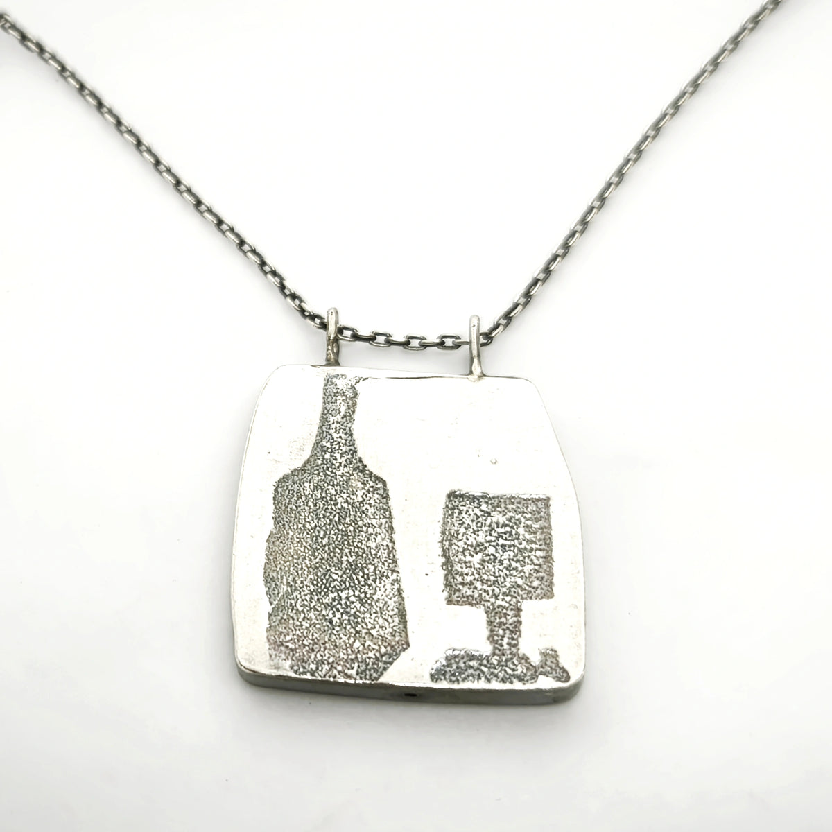 Close-up detail of a sterling silver pendant featuring a bottle and glass motif suspended from a silver chain, by Inês Almeida.