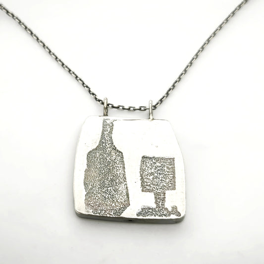 Close-up detail of a sterling silver pendant featuring a bottle and glass motif suspended from a silver chain, by Inês Almeida.