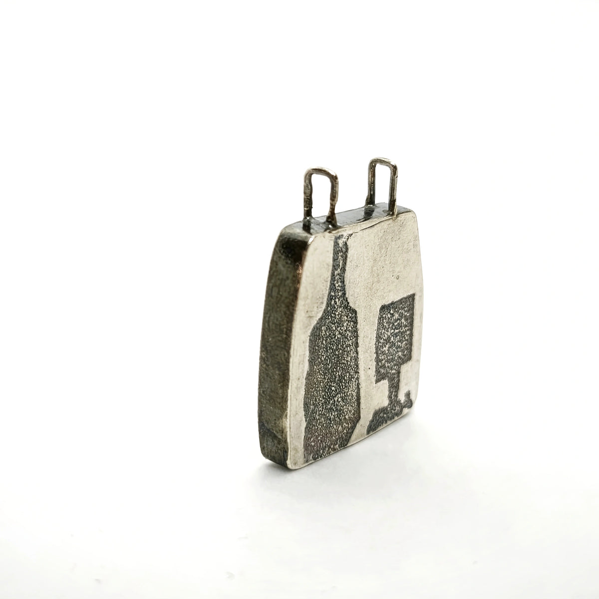 Profile view of a sterling silver pendant featuring a bottle and glass motif, showing its depth and suspension loops, by Inês Almeida.