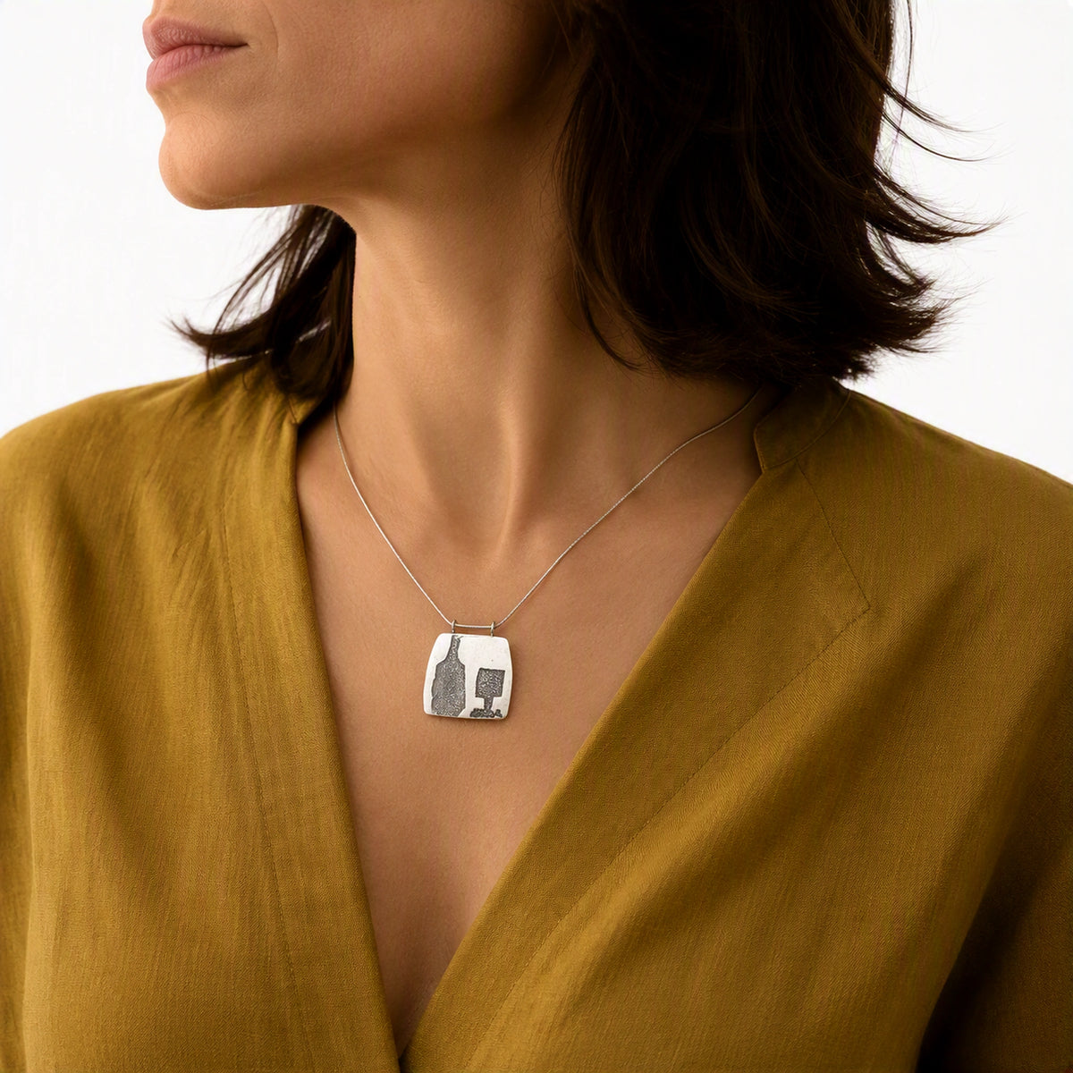 Sterling silver pendant featuring a bottle and glass motif worn on a contemporary mustard yellow garment, from the At the Table with Morandi collection by Inês Almeida.